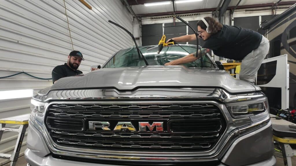 Two technicians performing simultaneous windshield tint shrinking and ceramic coating leveling on a silver Ram 1500 Limited inside the Englewood studio.